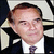 Static pixelated image of a person in formal attire with dark hair, light skin tone, and a neutral expression against a blurred background.