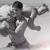 Black-and-white image of a shirtless man holding a woman in his arms, both in dynamic poses against a plain background.