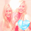 Two people with long blonde hair pose closely together, one wearing a blue top with a white strap, against a light background.