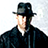 Static pixelated image of a person wearing a dark fedora and dark coat with light-colored shirt, set against a white background.