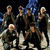 Group of people in dark clothing posed on a light surface with blue background, two versions show different cropping and pixelation levels.