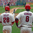 Two baseball players in white uniforms with red numbers 28 and 8, wearing red helmets, standing on a green field.