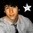 Pixelated image of a person in a light gray jacket with a black tie, standing against a black background with a white star symbol to the right.