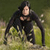 A pixelated figure in a black wetsuit stands in shallow water, arms extended, against a blurred greenish background.