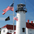 Static pixelated image of a white lighthouse with dark top, red-roofed buildings, and an American flag on a pole against a blue sky.