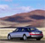 A static pixelated image shows a blue sedan with red accents driving on a road under a cloudy sky with distant hills.