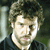Pixelated close-up of a person with light skin, dark curly hair, and a dark shirt against a blurred background.