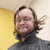A pixelated static image shows a person with long brown hair and a beard wearing a dark turtleneck sweater against a yellow and white background.