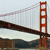 Static pixelated image of a red suspension bridge with white cables against a light gray sky, featuring a dark brown roadway and water below.
