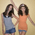 Two women stand side-by-side against a beige background; one wears a light blue sleeveless top and dark blue shorts, the other an orange sleeveless dress with blue trim.