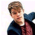 Static pixelated image of a person with light brown hair wearing dark blue clothing and red accents, cropped to show upper body against white background.