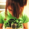 A person with brown hair in a high ponytail wears a green t-shirt with a black mushroom graphic and a small red heart, holding a silver digital camera.