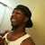 A person in a white tank top and dark cap smiles while turning their head left to right against a light-colored wall with a ceiling fixture.