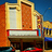 Pixelated image of a building facade with orange and beige architectural details, a white horizontal band, and blue sky background.