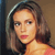 Pixelated close-up of a person with shoulder-length brown hair, wearing a dark top with a light-colored strap visible on the shoulder.