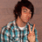 Pixelated image of a person with short dark hair wearing a blue and white checkered shirt, making a peace sign gesture against a brown background.