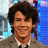 Pixelated image of a person with dark curly hair wearing a light brown suit jacket over a white shirt and black tie, set against a blue and gray background.
