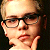 Pixelated close-up of a person with light hair and glasses, wearing dark clothing against a black background.