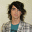 Pixelated portrait of a person with dark shoulder-length hair wearing a teal shirt and gray patterned jacket against a light beige background.
