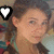 A pixelated image of a person with brown hair in pigtails wearing a teal necklace and dark top, with a white heart icon in the corner.