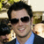 Pixelated image of a person wearing dark sunglasses, light blue shirt, and dark jacket with arm around another figure in background.