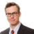 Pixelated portrait of a person with short brown hair, wearing dark-framed glasses, a white dress shirt, and a dark suit jacket with a red patterned tie against a light gray background.