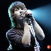 Static image of a person singing into a microphone on stage with blue lighting and dark background.