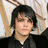 Pixelated static image of a person with dark hair wearing a dark jacket over a light shirt, set against a blurred background with light and brown tones.