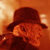 Static 50x50 pixel icon showing a dark brown fedora hat with wide brim and orange-red background, partially obscuring a blurred figure's face with visible eyes and mouth.