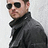 Pixelated static image of a person wearing dark sunglasses and a high-collared black jacket against a light gray background.
