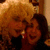 Two women with curly blonde and dark hair pose closely together, one wearing a red earring; green text appears in the bottom-right corner.