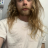 A pixelated static image shows a person with long brown hair wearing a white shirt against a light background.