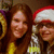 Two women with long brown hair pose together; one wears a red and white Santa hat, the other has no headwear.
