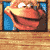 Pixelated character face with orange skin, white eyes, and a wide grin on blue background within wooden frame.