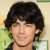 Pixelated close-up of a person with dark hair and light skin, wearing a dark top against a blurred green and beige background.