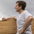 Pixelated image of a person in profile wearing a white shirt, standing beside a wooden structure against a light gray background.