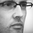 Pixelated grayscale close-up of a person's face wearing dark-framed glasses, showing eyes and nose with visible mouth area.