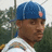 Pixelated image of a person wearing a blue cap with a pink emblem, white shirt, and dark background with green foliage.