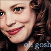 Close-up of a person with light brown curly hair, smiling with red lips, wearing a dark top with visible text 'oh gosh' in light blue.