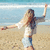 A woman with long blonde hair in a white crop top and light blue shorts stands on a sandy beach with arms outstretched, one arm raised high, against a blue ocean background.