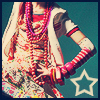 A person in traditional attire with layered red and pink beaded necklaces, a white shawl, and red-and-white striped arm coverings poses against a teal background with a white star outline.