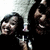 Pixelated static image shows two figures in dark clothing against a black background, one holding a light-colored object resembling a sword or staff.