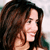A static pixelated image shows a person with long dark brown hair, light skin, and a slight smile, wearing a dark top against a light background.