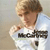 Pixelated image of a person with light brown hair wearing a white shirt with dark text, holding a small object near their chest.