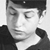 Black-and-white pixelated image of a person wearing a dark cap and dark clothing, with a hand near the face in a contemplative pose.