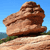 Pixelated image of a large brown rock formation on a light-colored base with green vegetation and blue sky background.