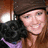 A pixelated image shows a person with long brown hair wearing a dark cap and purple clothing, holding a black dog against a blurred background.