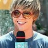 A person with short light brown hair wearing large brown sunglasses and a blue shirt holds a black microphone with red accents.