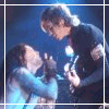 Two people in dark clothing and light blue sleeves lean toward each other under stage lighting, with text reading Angels in Everyone below.