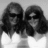 Two pixelated figures with long dark hair and white clothing against a light gray background, rendered in grayscale.